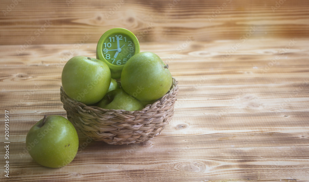 The green apple and green alarm clock in woven basket ,put on wooden timber boardvintage warm light tone,blurry light around. The green apple and green alarm clock in woven basket ,put on wooden timber boardvintage warm light tone,blurry light around.