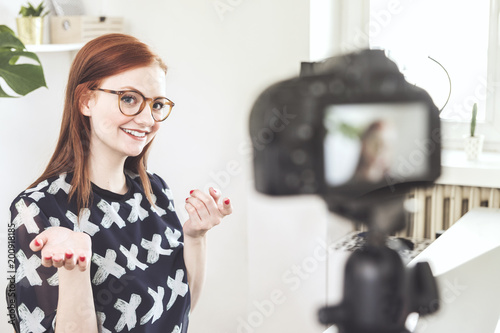 Young  redhead female blogger recording video at home using camera with a tripod.