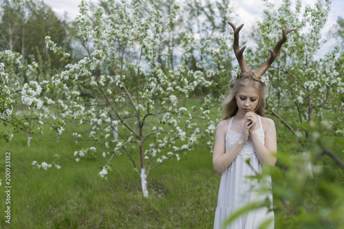 Teen beautiful blonde girl wearing white dress with deer horns on her head and white flowers in hair stays in a spring blooming garden of trees covered with white flowers.