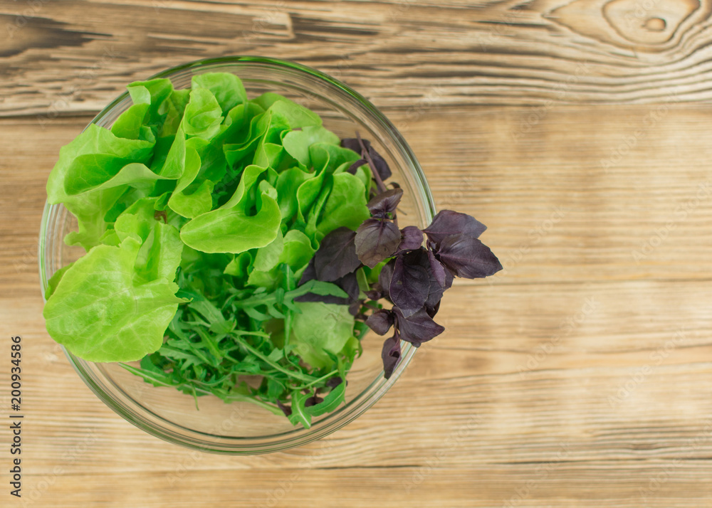 Fresh lettuce leaves, basil, arugula in a glass bowl on a wooden background. Preparation of a vegetarian vegetable salad.