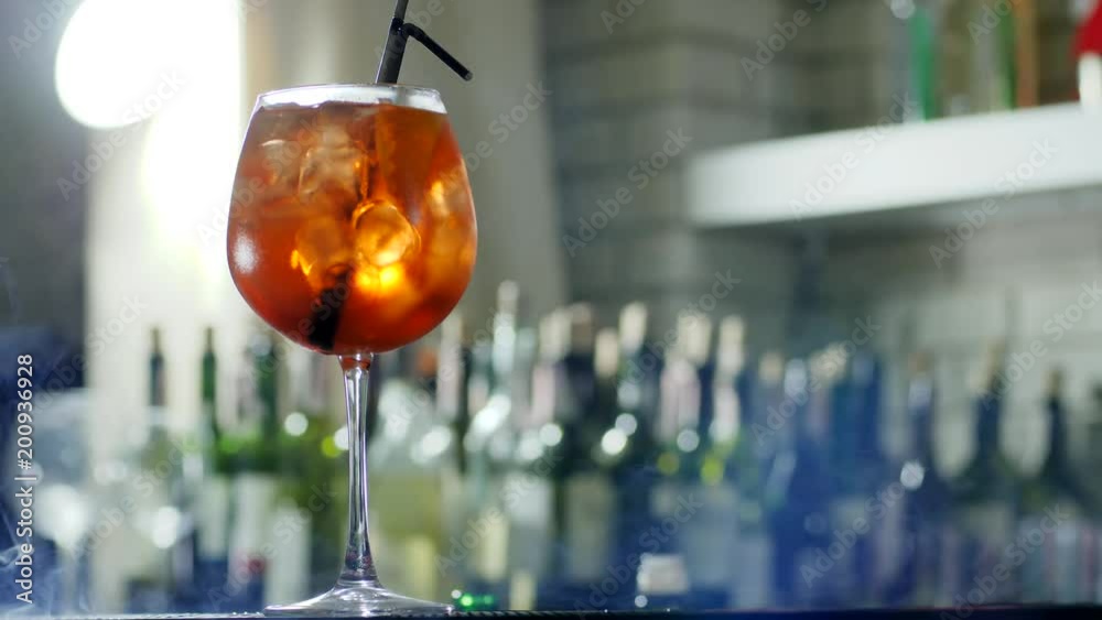 bright alcohol drink with ice cubes and plastic straw in wineglass closeup into easy haze on unfocused background of bottles