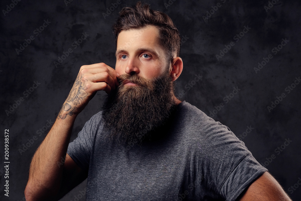 Portrait of a hipster with full beard and stylish haircut, dressed in a gray t-shirt, stands with a thinking look in a studio on a dark background. Portrait of a hipster with full beard and stylish haircut, dressed in a gray t-shirt, stands with a thinking look in a studio on a dark background.
