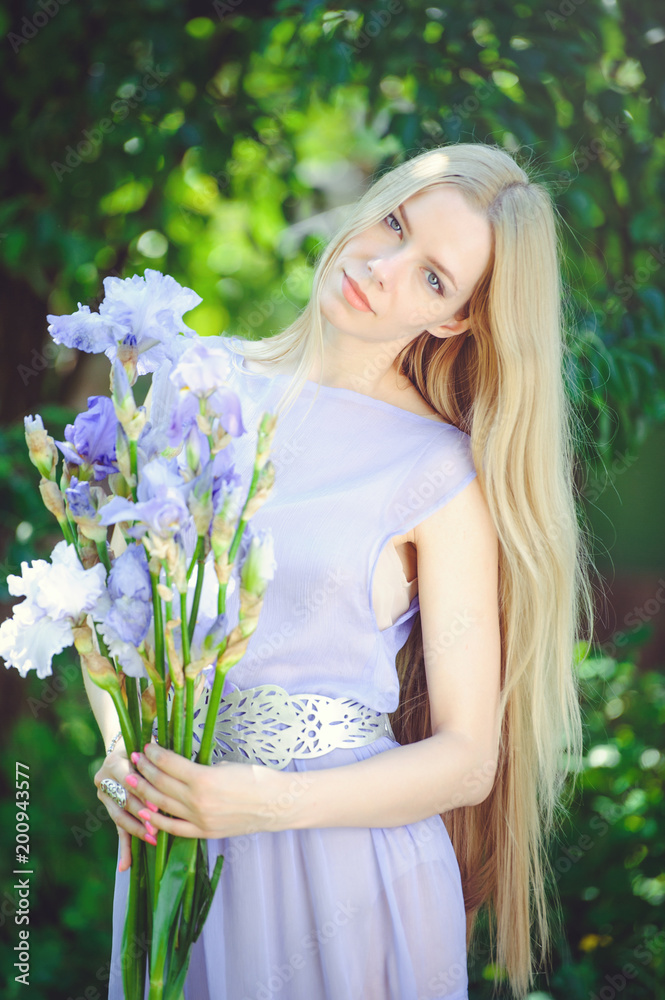 Attractive young girl with blonde dren hair and natural make-up smelling blue purple iris flowers on a background outdoors, tenderness and softness on a nature background Attractive young girl with blonde dren hair and natural make-up smelling blue purple iris flowers on a background outdoors, tenderness and softness on a nature background