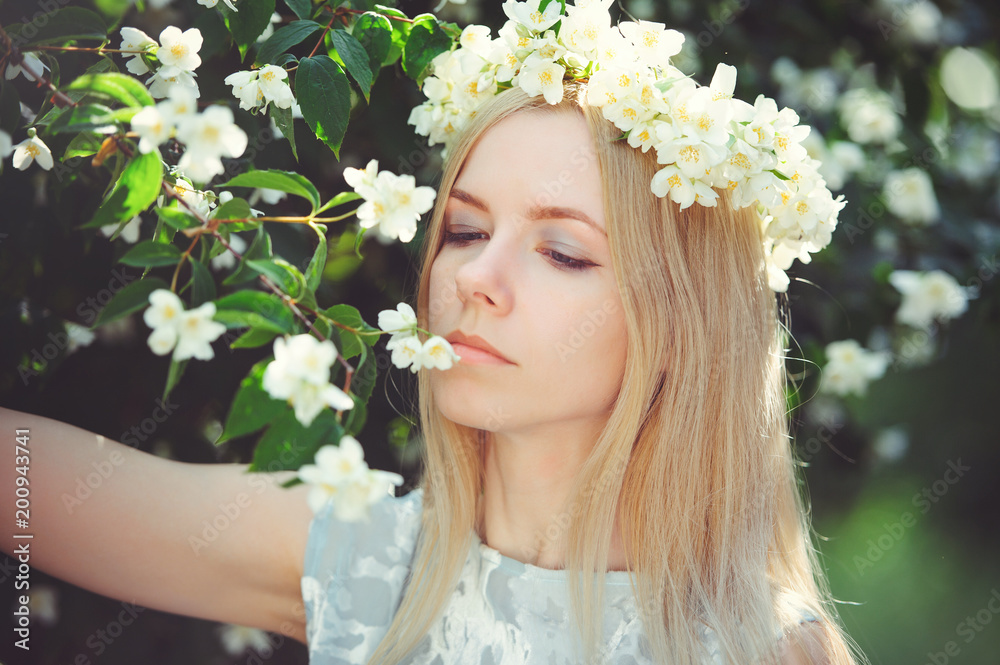 Fototapeta premium Attractive modest young girl with blonde with jasmine flowers wreath on head long hair and natural make-up in white dress outdoors, tenderness and softness on nature background