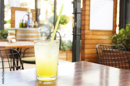 A glass of fresh ice cold lemonade on wooden table of coffee shop patio with green bushes on background. Close up of refreshing lemon juice cold beverage on the cafe terrace, outside. Text copy space