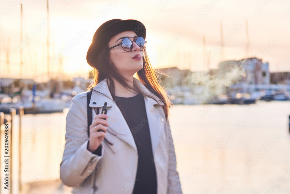 Fototapeta premium Pretty young woman in black hat with vape at a sea port