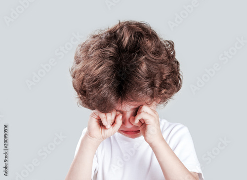 Cute little curly-headed boy in a white t-shirt is crying and looking at the camera. Gray background. Close-up.