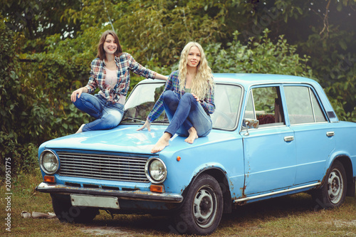 cheerful Attractive two young blonde girl and brunette posing on the hood of an old rusty car, dressed in jeans and shirts on a nature background