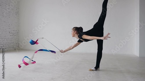 Little gymnastics in black body doing the gymnastics exercise with a colored ribbon in the white studio. Cute girl makes stretching exercise in sport gym photo