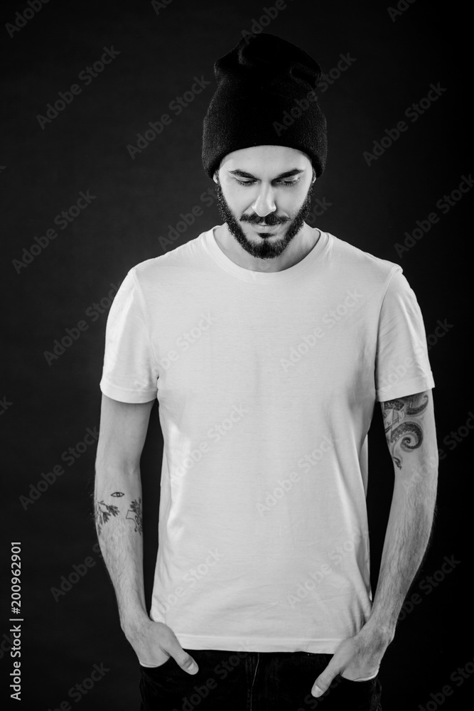 Waist up portrait of serious stylish attractive man with thick beard looks confidently, dressed in casual white t shirt, isolated over black background. Young hipster in cap with tattoo. Mock up Waist up portrait of serious stylish attractive man with thick beard looks confidently, dressed in casual white t shirt, isolated over black background. Young hipster in cap with tattoo. Mock up