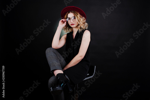 Studio portrait of blonde girl in black wear, red hat and glasses against dark background.