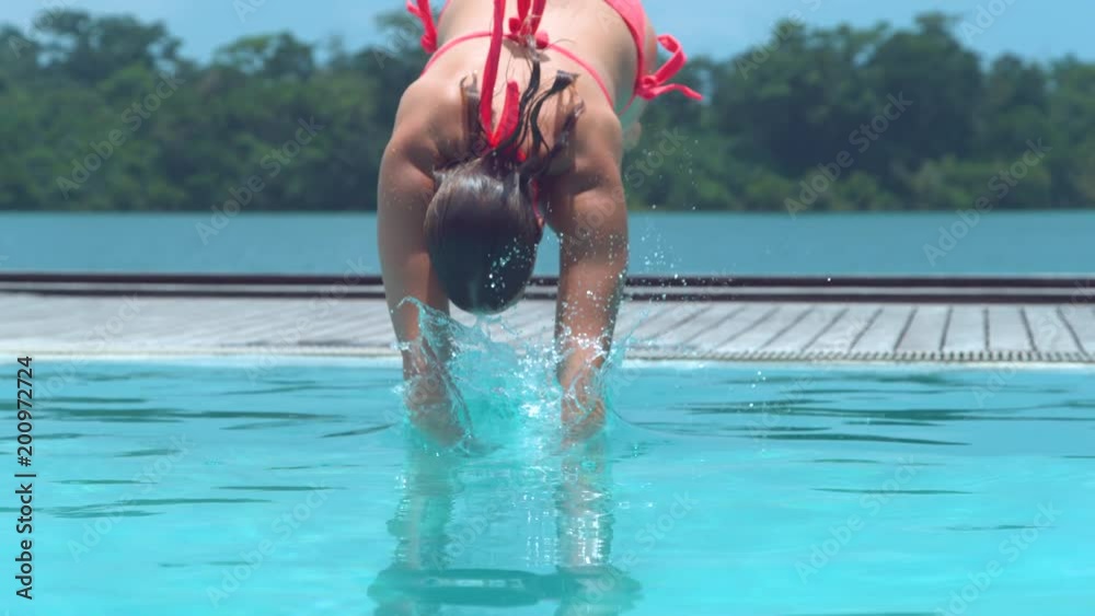 SLOW MOTION, CLOSE UP: Carefree female traveler jumps into cool beachfront pool in Vanuatu during her relaxing summer vacation. Young Caucasian woman dives into empty pool and splashes glassy water.