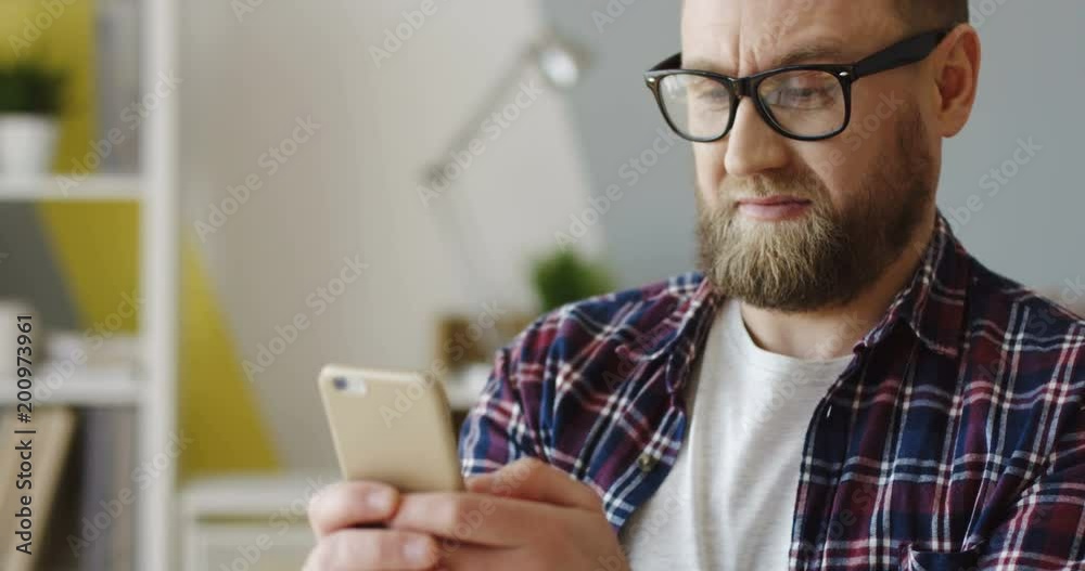 Close up of the Caucasian man with a beard in glasses and jeans shirt holding a smartphone and taping on it with emotions. Indoors Close up of the Caucasian man with a beard in glasses and jeans shirt holding a smartphone and taping on it with emotions. Indoors