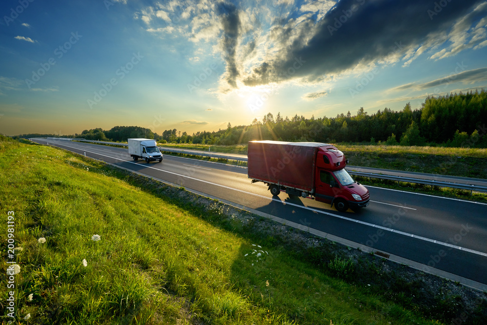 Red and white delivery van driving on the highway in the countryside in the rays of the sunset with dramatic clouds