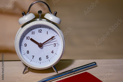 front view of white clock with pen and red notebook on the wood table