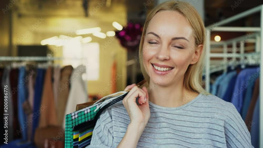Close-up portrait of blond girl standing with paper bags in clothing boutique, laughing and smiling happily and looking at camera. Stylish clothes is in background. Close-up portrait of blond girl standing with paper bags in clothing boutique, laughing and smiling happily and looking at camera. Stylish clothes is in background.