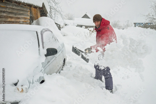 a man in a red jacket cleans the snow near his gray car to release his snow blockage