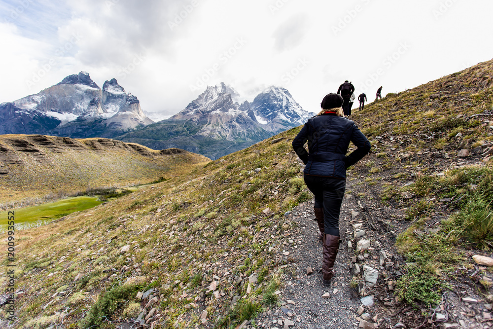 woman dressed in dark clothing walking along pathway with Torres del Paine mountain range in the back - obrazy, fototapety, plakaty 