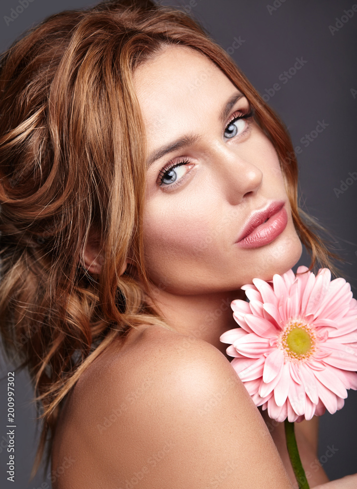 Beauty fashion portrait of young blond woman model with natural makeup and perfect skin with bright pink gerbera flower posing in studio.