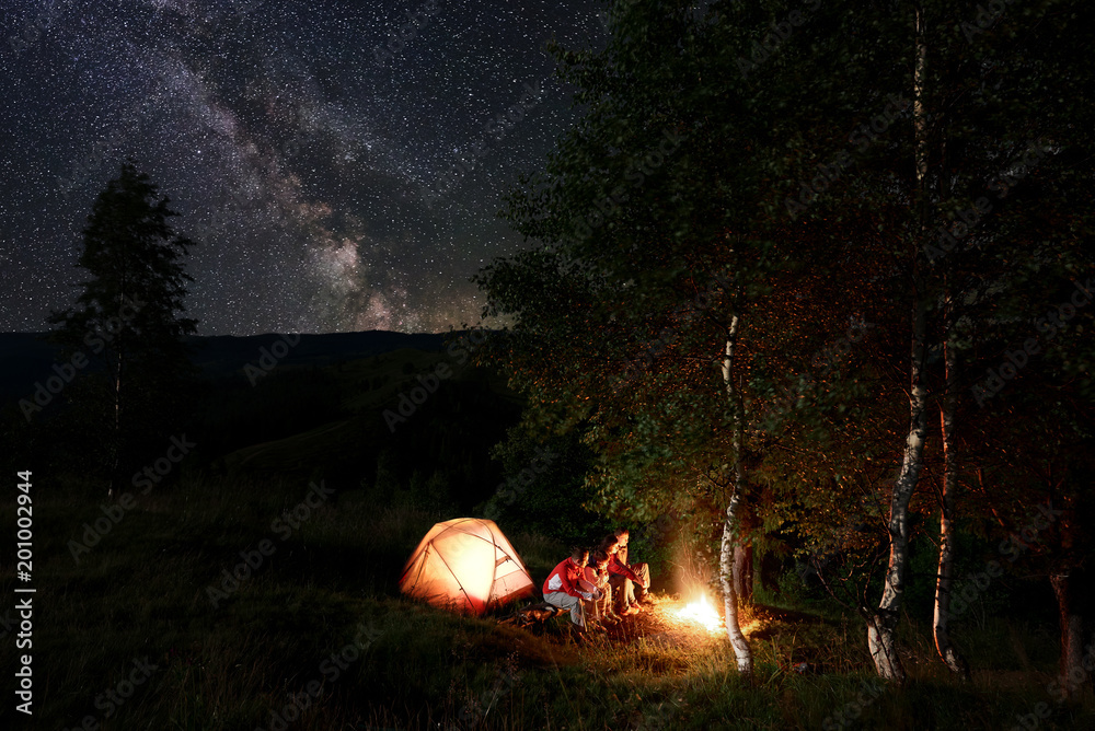 Plakat Four friends tourists having a rest by the campfire, sitting on logs during night camping among trees near the illuminated tent in the mountains under incredible beautiful starry sky with Milky way