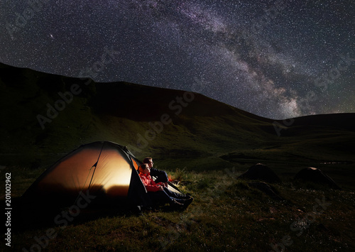 Man and woman having a rest at night camping in the Carpathians mountains near Dogyaska lake, sitting in tent, looking at the sky strewn with stars. In the tent lights on. Concept of active recreation