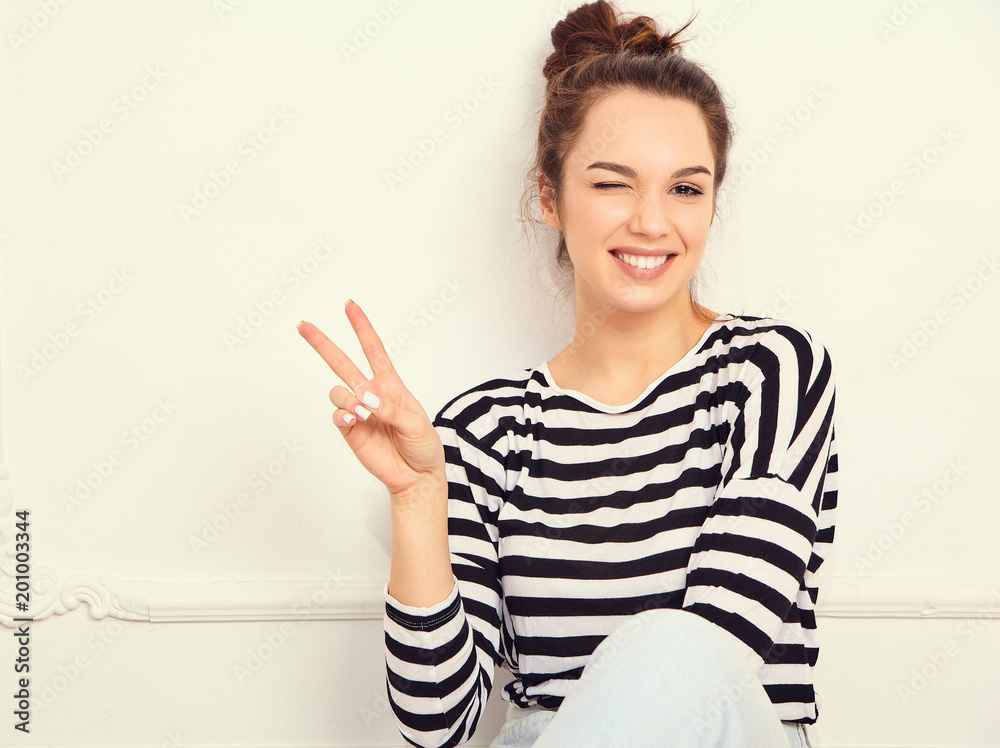 Portrait of young beautiful brunette woman girl model with nude makeup in summer hipster clothes posing near wall. Showing peace sign and winking Portrait of young beautiful brunette woman girl model with nude makeup in summer hipster clothes posing near wall. Showing peace sign and winking