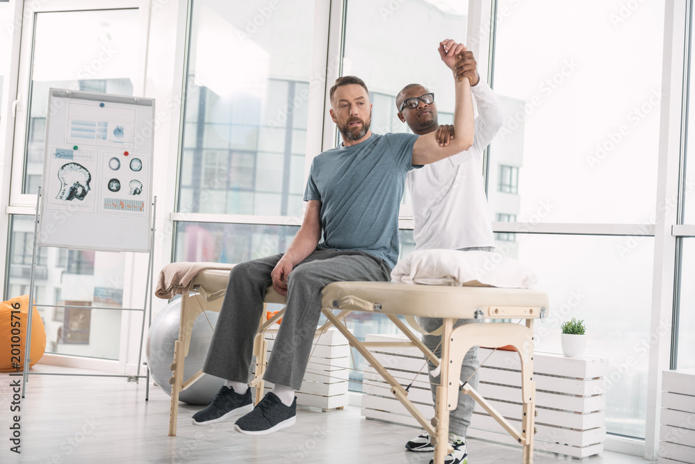 Medical rehabilitation. Skillful nice doctor raising his patients arm while doing medical rehabilitation Medical rehabilitation. Skillful nice doctor raising his patients arm while doing medical rehabilitation
