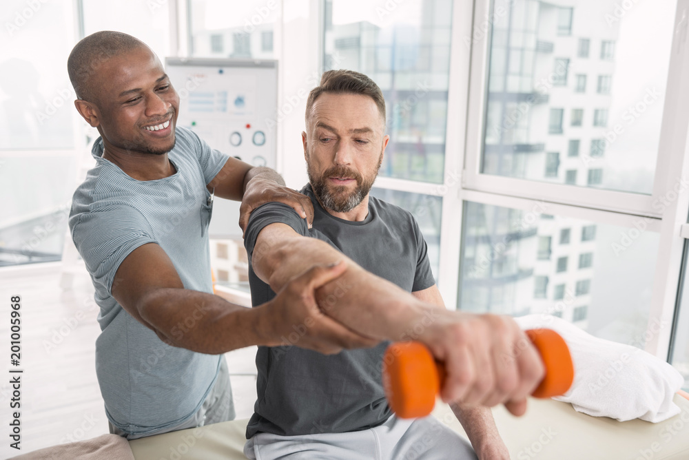 Fototapeta premium Favourite job. Delighted nice therapist smiling to his patient while helping his to lift a hand