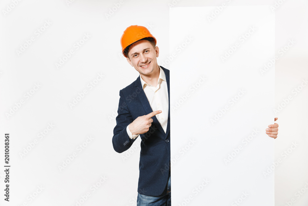 Smiling businessman in dark suit, protective hardhat holding big white empty blank material with place for text or image and pointing index finger aside on copy space isolated on white background. Smiling businessman in dark suit, protective hardhat holding big white empty blank material with place for text or image and pointing index finger aside on copy space isolated on white background.