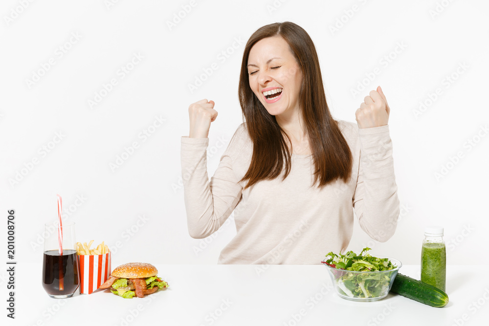 Overjoyed woman at table with green detox smoothies, salad in glass bowl, cucumber, burger, cola in bottle isolated on white background. Proper nutrition, healthy lifestyle, fast food, dieting concept Overjoyed woman at table with green detox smoothies, salad in glass bowl, cucumber, burger, cola in bottle isolated on white background. Proper nutrition, healthy lifestyle, fast food, dieting concept