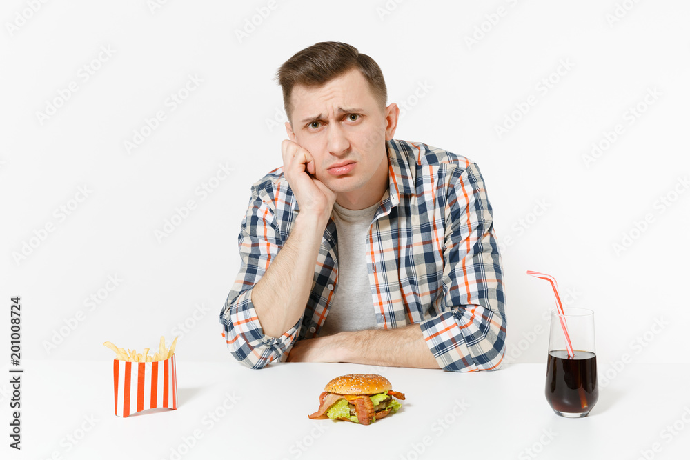 Handsome sad upset young man sitting at table with burger, french fries, cola in glass isolated on white background. Proper nutrition or American classic fast food. Advertising area with copy space. Handsome sad upset young man sitting at table with burger, french fries, cola in glass isolated on white background. Proper nutrition or American classic fast food. Advertising area with copy space.