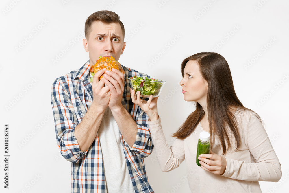 Couple man and woman standing with green detox smoothies in bottle, salad in glass bowl, burger isolated on white background. Proper nutrition, healthy lifestyle, fast food, choice concept. Copy space Couple man and woman standing with green detox smoothies in bottle, salad in glass bowl, burger isolated on white background. Proper nutrition, healthy lifestyle, fast food, choice concept. Copy space
