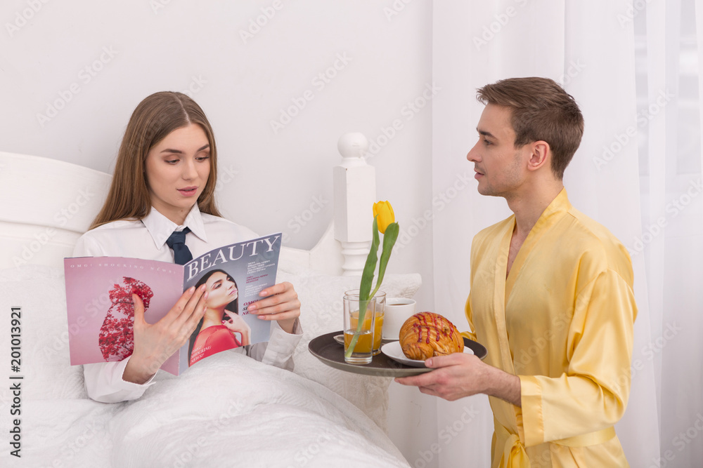 Your servant. Dominant concentrated woman reading magazine and her boyfriend bringing her breakfast in bed Your servant. Dominant concentrated woman reading magazine and her boyfriend bringing her breakfast in bed