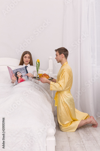Making your happy. Discontent concentrated woman reading magazine and her boyfriend bringing her breakfast in bedtisfied businesswoman reading a magazine
