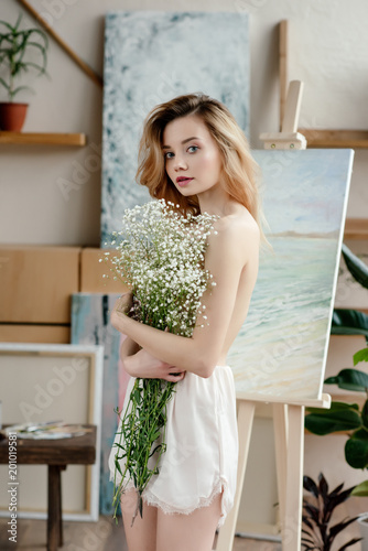 beautiful naked young woman holding white flowers and looking at camera in art studio