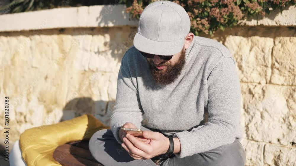 Grown man wearing grey sweater, jeans, cap and shandes sits on a bench outdoor under the sun. He is holding smartphone and chatting. Grown man wearing grey sweater, jeans, cap and shandes sits on a bench outdoor under the sun. He is holding smartphone and chatting.