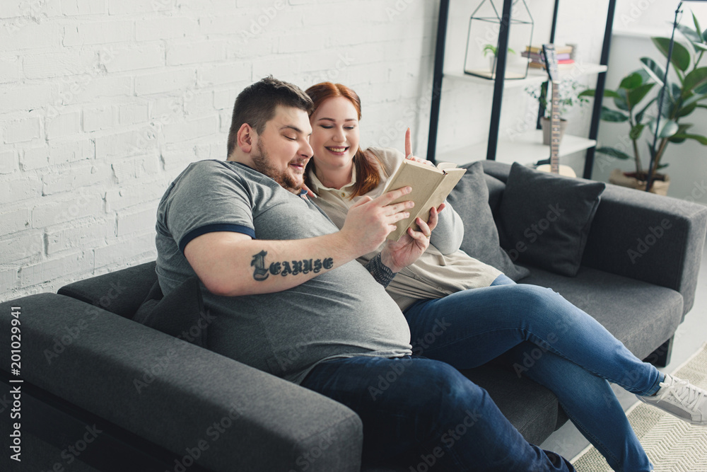 Obraz premium Smiling overweight boyfriend and girlfriend reading book on sofa at home