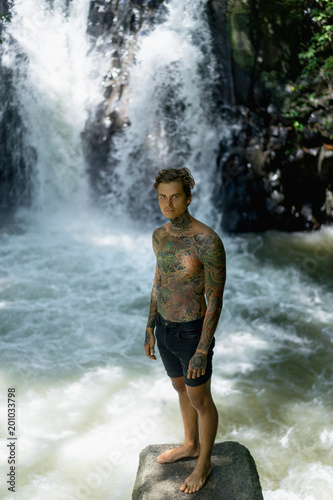tattooed man standing on rock with Aling-Aling Waterfall on background, Bali, Indonesia
