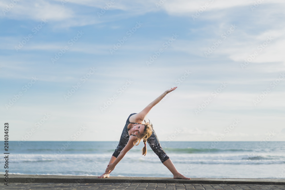 woman in Extended Triangle yoga pose with ocean and blue sky on background, Bali, Indonesia