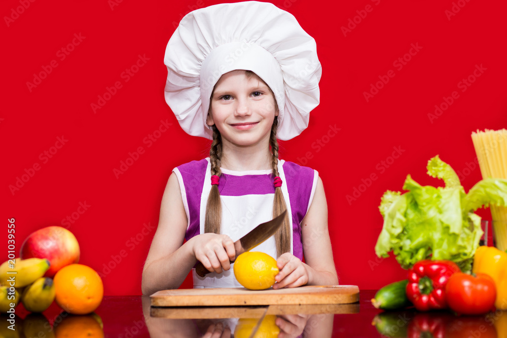 Happy little girl in chef uniform cuts fruit in kitchen. Kid chef. Cooking Process Concept Happy little girl in chef uniform cuts fruit in kitchen. Kid chef. Cooking Process Concept