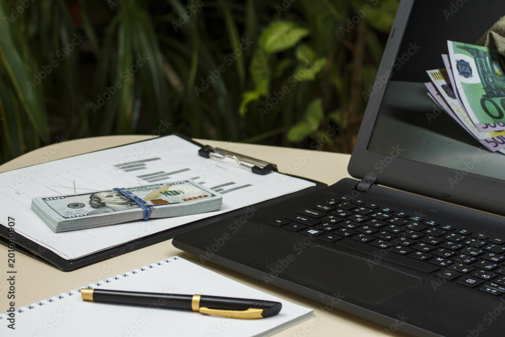 Laptop with dollars and telephone on the table on the background of plants Laptop with dollars and telephone on the table on the background of plants