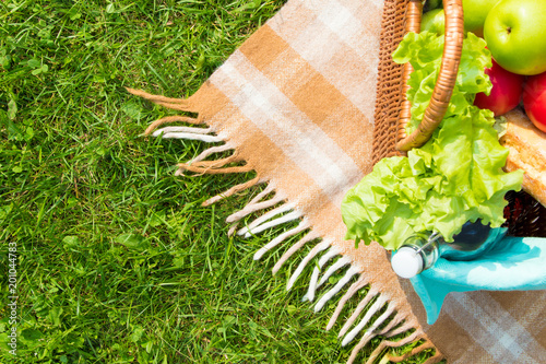 Green grass and straw basket with food and drinks on checkered beige tablecloth background for picnic, top view