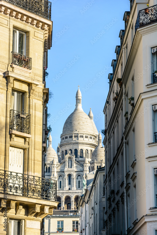 View of the facade and dome of the Basilica of the Sacred Heart in Paris through a narrow street between typical buildings under a clear blue sky.