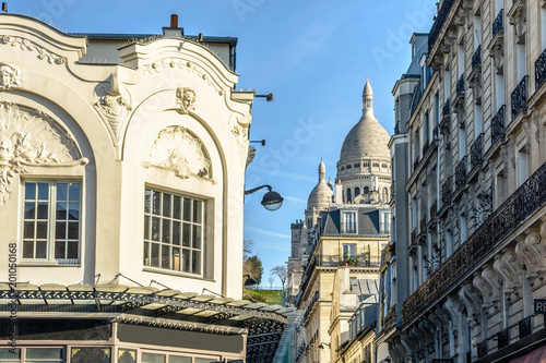 View of the facade and dome of the Basilica of the Sacred Heart of Paris through a narrow street between old parisian buildings under a clear blue sky.