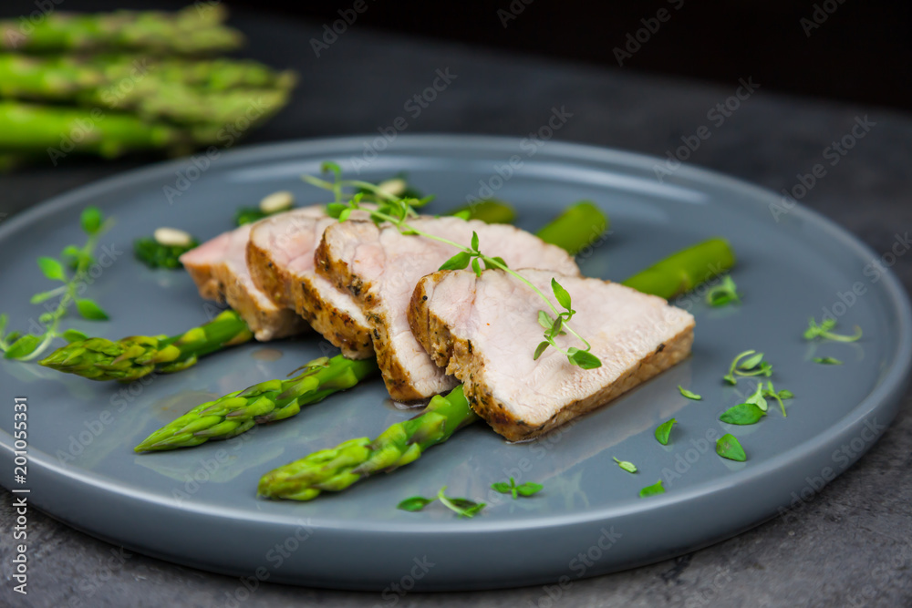 Slices of roasted pork tenderloin on asparagus with pesto from kale and thyme on the grey plate. A bunch of asparagus on the background Slices of roasted pork tenderloin on asparagus with pesto from kale and thyme on the grey plate. A bunch of asparagus on the background