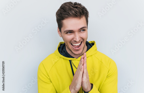 Studio closeup shot of sneaky sly and scheming young European man dressed in stylish yellow hoodie looking at camera with mysterious smiles, male making gesture as if washing his hands. Body language