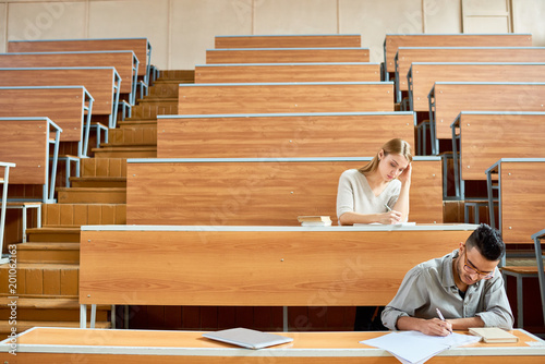 Low angle view of two students, one of them Middle-eastern sitting at desks in steep modern lecture hall at college and reading notes, copy space