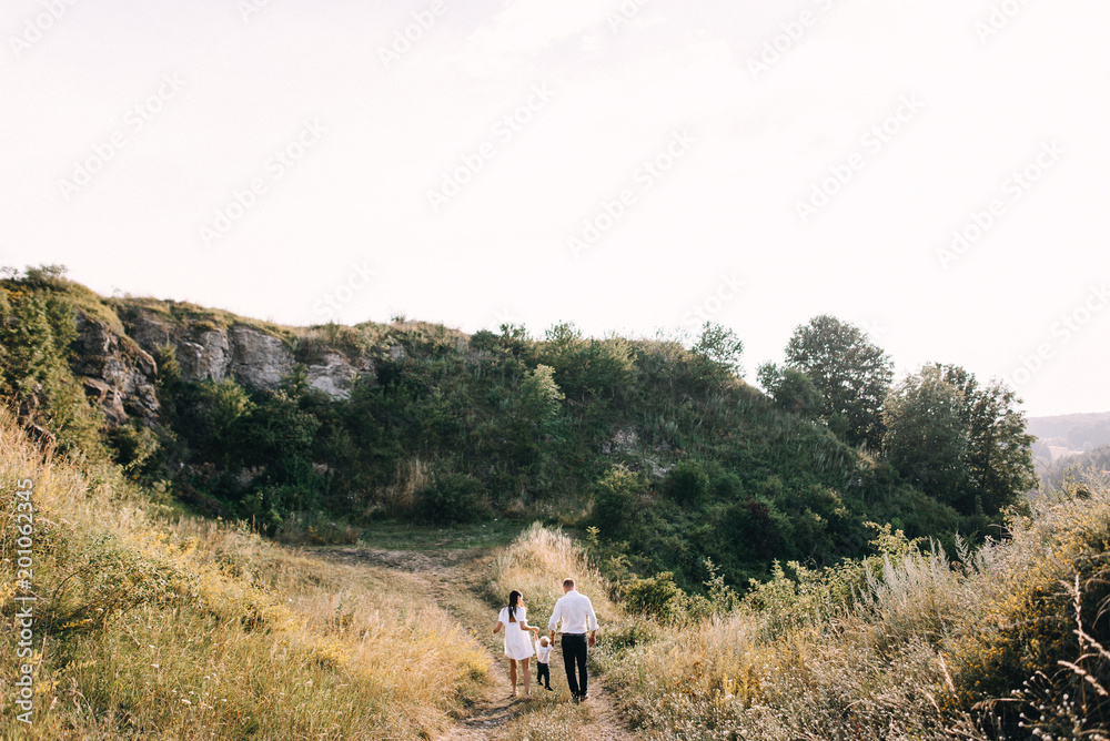 Walk beautiful young family in white clothes with a young son blond in mountainous areas with tall grass at sunset. Mother keeps son in her arms, hugging. family - this is happiness