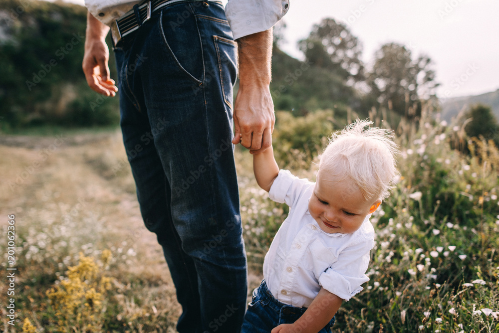 Fototapeta premium Walk beautiful young family in white clothes with a young son blond in mountainous areas with tall grass at sunset. portrait of a smiling boy