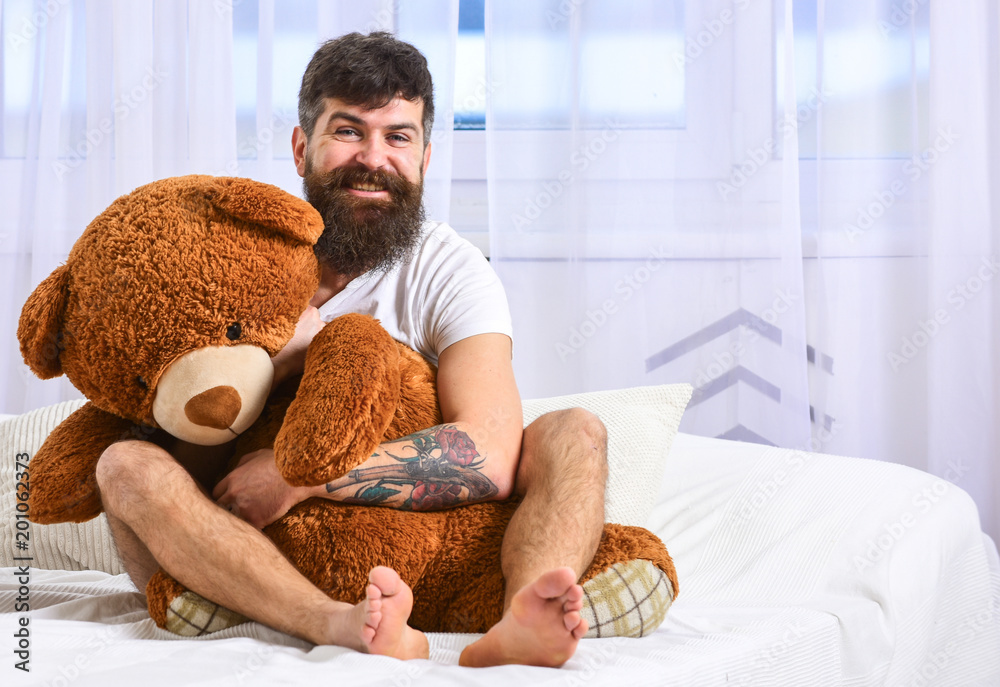 Obraz premium Guy on cheerful face hugs giant teddy bear. Infantilism concept. Macho with beard and mustache relaxing with plush toy after nap, rest. Man sits on bed and hugs big toy, white curtains on background.